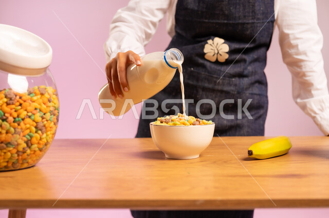 وجبة طعام صحية، تحضير رقائق الذرة في طبق أبيض، سكب الحليب فوق حبوب الإفطار، اتباع نظام غذائي صحي، بورتريه مقرب لامرأة عربية خليجية سعودية ترتدي اليونفر تقوم بإعداد الفطور الصباحي على طاولة المطبخ، خلفية ملونة