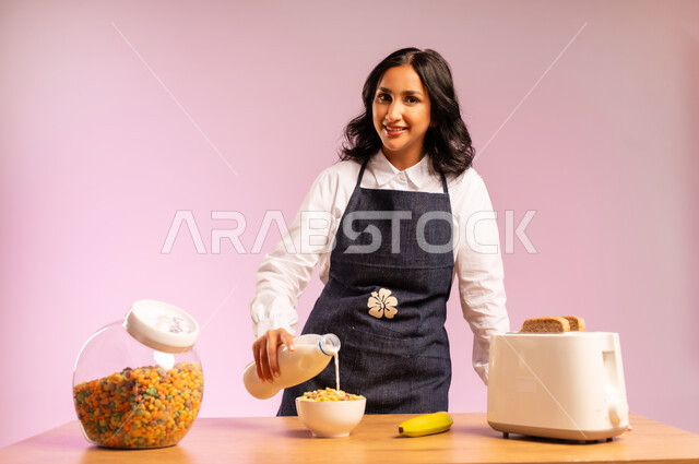 تحضير رقائق الذرة في طبق أبيض بايماءات السعادة والسرور، سكب الحليب فوق حبوب الإفطار، اتباع نظام غذائي صحي، بورتريه مقرب لامرأة عربية خليجية سعودية ترتدي اليونفر تقوم بإعداد الفطور الصباحي على طاولة المطبخ،  وجبة طعام صحية، خلفية ملونة