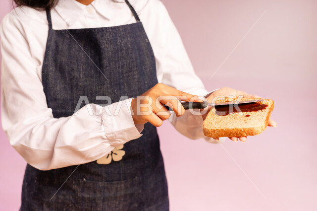 تحضير شرائح خبز التوست، ايماءات السعادة والسرور، اتباع نظام غذائي، بورتريه مقرب لامرأة عربية خليجية سعودية ترتدي اليونفر تقوم بإعداد الفطور الصباحي على طاولة المطبخ، مفهوم الأكل الصحي، وجبة طعام صحية، خلفية ملونة