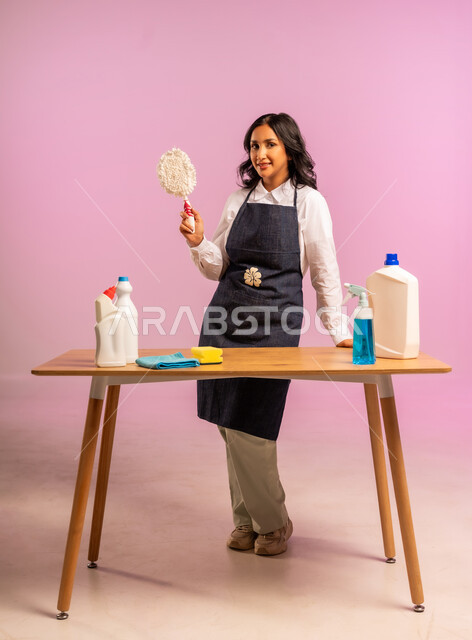 المهام والأعمال المنزلية الروتينية، التطهير والتنظيف في المنازل,  بورتريه لمرأة عربية خليجية سعودية ترتدي اليونفور وتحمل بيدها مواد ومستلزمات التعقيم، أداء مهام المطبخ، الجسم بالطول الكامل، خلفية ملونة