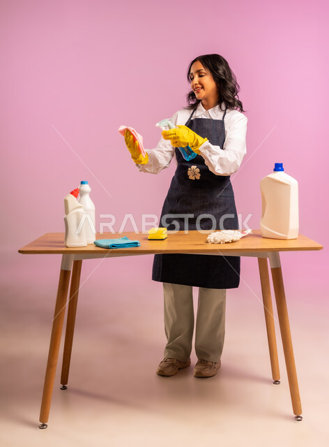 المهام والأعمال المنزلية الروتينية، التطهير والتنظيف في المنازل,  بورتريه لمرأة عربية خليجية سعودية ترتدي القفازات واليونفور وتحمل بيدها مواد ومستلزمات التعقيم، أداء مهام المطبخ، الجسم بالطول الكامل، خلفية ملونة