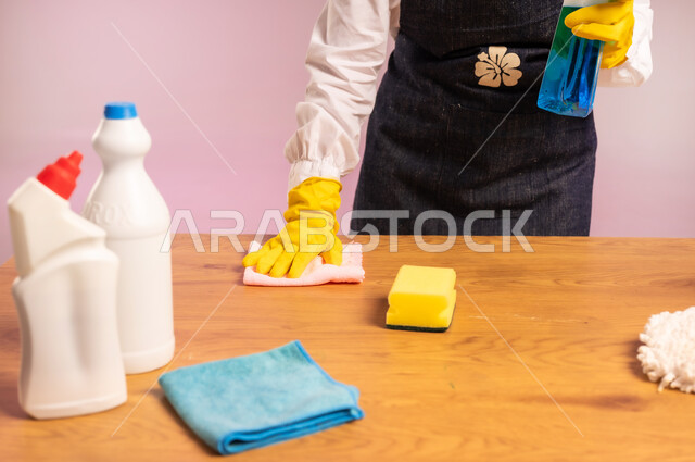 القيام بتنظيف طاولة خشبية، التطهير في المنازل, المهام والأعمال المنزلية الروتينية،  بورتريه مقرب ليدين امرأة عربية خليجية سعودية ترتدي القفازات واليونفور، أداء مهام المطبخ، الجسم بالطول الكامل، خلفية ملونة