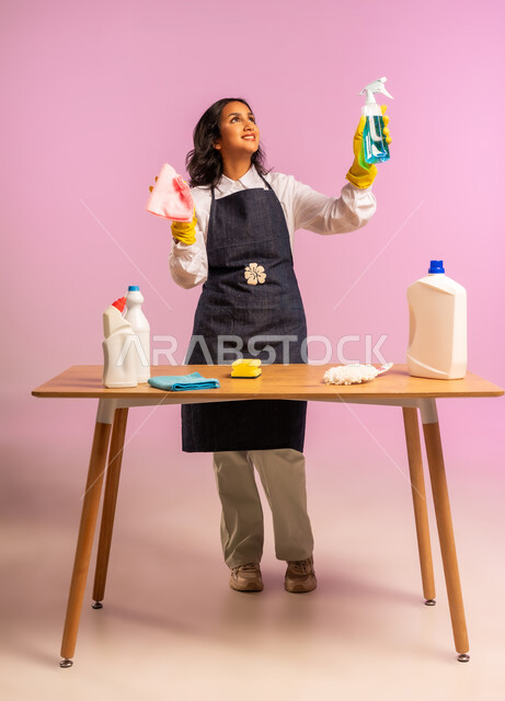 المهام والأعمال المنزلية الروتينية، التطهير والتنظيف في المنازل,  بورتريه لمرأة عربية خليجية سعودية ترتدي القفازات واليونفور وتحمل بيدها مواد ومستلزمات التعقيم، أداء مهام المطبخ، الجسم بالطول الكامل، خلفية ملونة