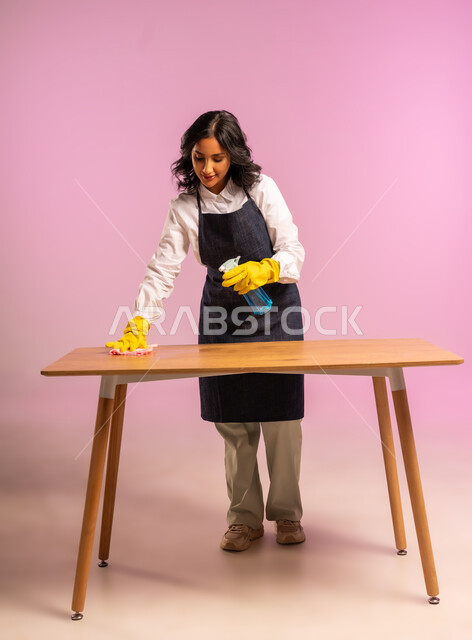 المهام والأعمال المنزلية الروتينية، التطهير والتنظيف في المنازل,  بورتريه لمرأة عربية خليجية سعودية ترتدي القفازات واليونفور وتحمل بيدها مواد ومستلزمات التعقيم، أداء مهام المطبخ، الجسم بالطول الكامل، خلفية ملونة
