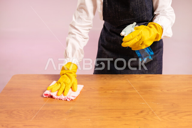 Cleaning and disinfection in homes, chores and routine household tasks, close-up portrait of hands of a Saudi Arabian Gulf woman wearing gloves and scrubber cleaning a wooden table with a sponge, performing kitchen tasks, full body, colorful background