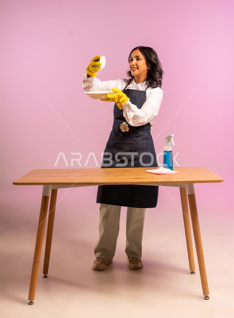 المهام والأعمال المنزلية الروتينية، القيام بغسل الأطباق بإسفنج الجلي وجل المنظف، بورتريه لسيدة عربية خليجية سعودية ترتدي القفازات واليونفور، النظافة وتطهير الادوات، أداء مهام المطبخ، الجسم بالطول الكامل، خلفية ملونة