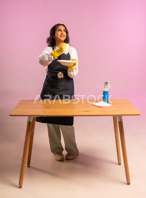 المهام والأعمال المنزلية الروتينية، القيام بغسل الأطباق بإسفنج الجلي وجل المنظف، بورتريه لسيدة عربية خليجية سعودية ترتدي القفازات واليونفور، النظافة وتطهير الادوات، أداء مهام المطبخ، الجسم بالطول الكامل، خلفية ملونة