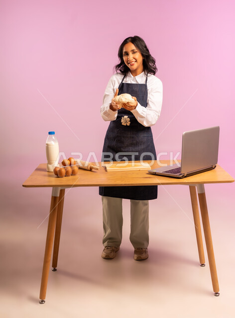 إستخدام أداة فرد العجين اليد (الشوبك)، بورتريه لمرأة عربية خليجية سعودية ترتدي اليونفور و تقوم بإعداد المخبوزات العربية، مشاركة تحضير الوصفات على مواقع التواصل الإجتماعي بإستخدام جهاز الحاسب المحمول، الجسم بالطول الكامل، خلفية ملونة