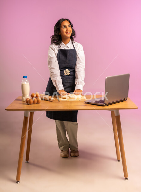 إستخدام أداة فرد العجين اليد (الشوبك)، بورتريه لمرأة عربية خليجية سعودية ترتدي اليونفور و تقوم بإعداد المخبوزات العربية، مشاركة تحضير الوصفات على مواقع التواصل الإجتماعي بإستخدام جهاز الحاسب المحمول، الجسم بالطول الكامل، خلفية ملونة