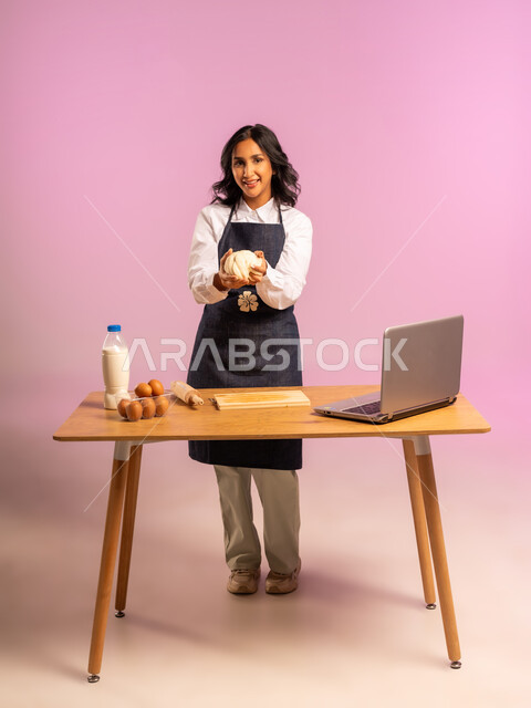 مشاركة تحضير الوصفات على مواقع التواصل الإجتماعي بإستخدام جهاز الحاسب المحمول، إستخدام أداة فرد العجين اليد (الشوبك)، بورتريه لمرأة عربية خليجية سعودية ترتدي اليونفور و تقوم بإعداد المخبوزات العربية، الجسم بالطول الكامل، خلفية ملونة