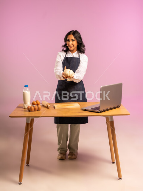 إستخدام أداة فرد العجين اليد (الشوبك)، مشاركة تحضير الوصفات على مواقع التواصل الإجتماعي بإستخدام جهاز الحاسب المحمول، بورتريه لمرأة عربية خليجية سعودية ترتدي اليونفور و تقوم بإعداد المخبوزات العربية، الجسم بالطول الكامل، خلفية ملونة
