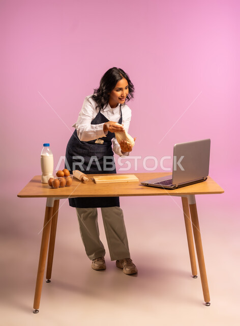 مشاركة تحضير الوصفات على مواقع التواصل الإجتماعي بإستخدام جهاز الحاسب المحمول، إستخدام أداة فرد العجين اليد (الشوبك)، بورتريه لمرأة عربية خليجية سعودية ترتدي اليونفور و تقوم بإعداد المخبوزات العربية، الجسم بالطول الكامل، خلفية ملونة