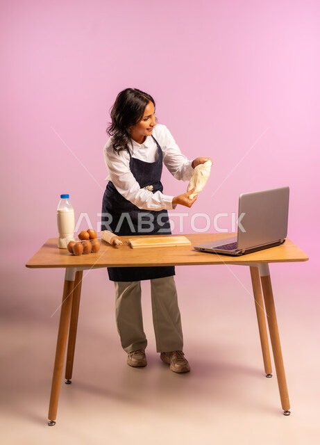 إستخدام أداة فرد العجين اليد (الشوبك)، بورتريه لمرأة عربية خليجية سعودية ترتدي اليونفور و تقوم بإعداد المخبوزات العربية، مشاركة تحضير الوصفات على مواقع التواصل الإجتماعي بإستخدام جهاز الحاسب المحمول، الجسم بالطول الكامل، خلفية ملونة