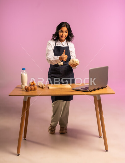 إستخدام أداة فرد العجين اليد (الشوبك)، مشاركة تحضير الوصفات على مواقع التواصل الإجتماعي بإستخدام جهاز الحاسب المحمول، بورتريه لمرأة عربية خليجية سعودية ترتدي اليونفور و تقوم بإعداد المخبوزات العربية، الجسم بالطول الكامل، خلفية ملونة