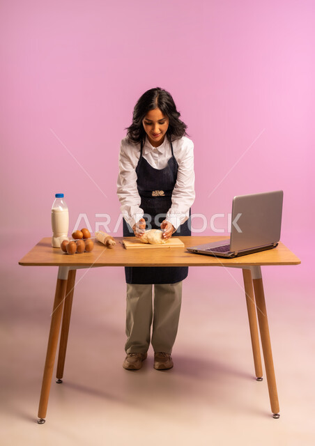 مشاركة تحضير الوصفات على مواقع التواصل الإجتماعي بإستخدام جهاز الحاسب المحمول، إستخدام أداة فرد العجين اليد (الشوبك)، بورتريه لمرأة عربية خليجية سعودية ترتدي اليونفور و تقوم بإعداد المخبوزات العربية، الجسم بالطول الكامل، خلفية ملونة