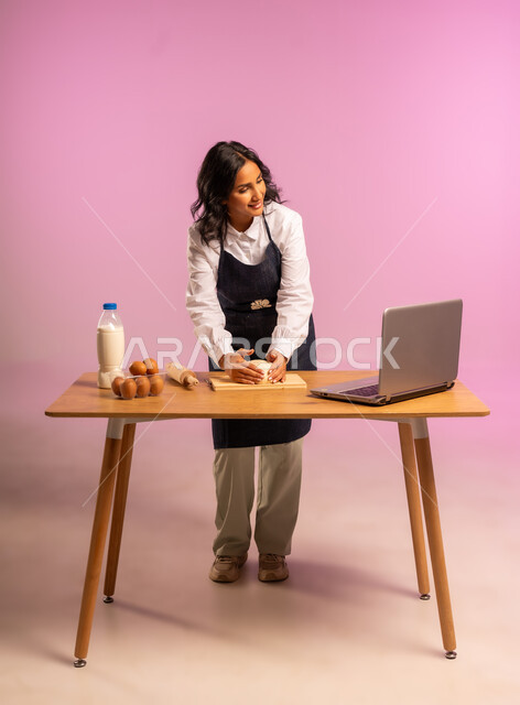 إستخدام أداة فرد العجين اليد (الشوبك)، بورتريه لمرأة عربية خليجية سعودية ترتدي اليونفور و تقوم بإعداد المخبوزات العربية، مشاركة تحضير الوصفات على مواقع التواصل الإجتماعي بإستخدام جهاز الحاسب المحمول، الجسم بالطول الكامل، خلفية ملونة