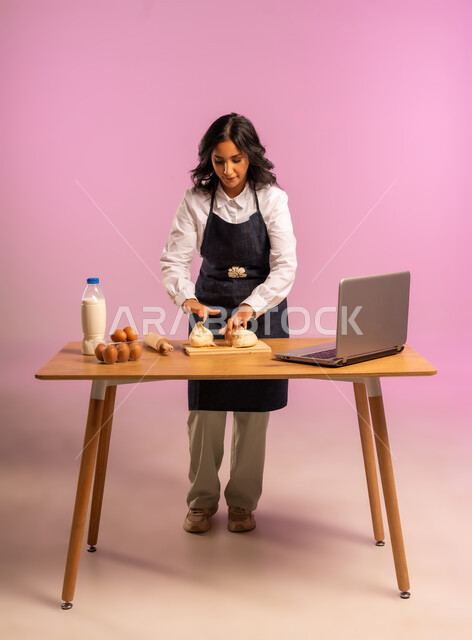 مشاركة تحضير الوصفات على مواقع التواصل الإجتماعي بإستخدام جهاز الحاسب المحمول، إستخدام أداة فرد العجين اليد (الشوبك)، بورتريه لمرأة عربية خليجية سعودية ترتدي اليونفور و تقوم بإعداد المخبوزات العربية، الجسم بالطول الكامل، خلفية ملونة