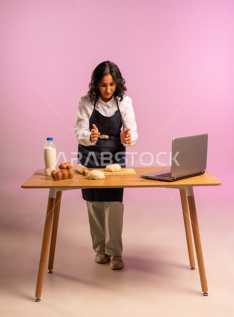 مشاركة تحضير الوصفات على مواقع التواصل الإجتماعي بإستخدام جهاز الحاسب المحمول، إستخدام أداة فرد العجين اليد (الشوبك)، بورتريه لمرأة عربية خليجية سعودية ترتدي اليونفور و تقوم بإعداد المخبوزات العربية، الجسم بالطول الكامل، خلفية ملونة