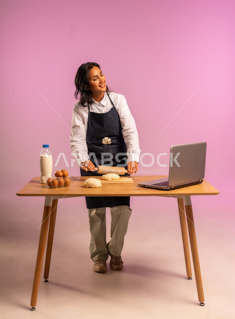 مشاركة تحضير الوصفات على مواقع التواصل الإجتماعي بإستخدام جهاز الحاسب المحمول، إستخدام أداة فرد العجين اليد (الشوبك)، بورتريه لمرأة عربية خليجية سعودية ترتدي اليونفور و تقوم بإعداد المخبوزات العربية، الجسم بالطول الكامل، خلفية ملونة