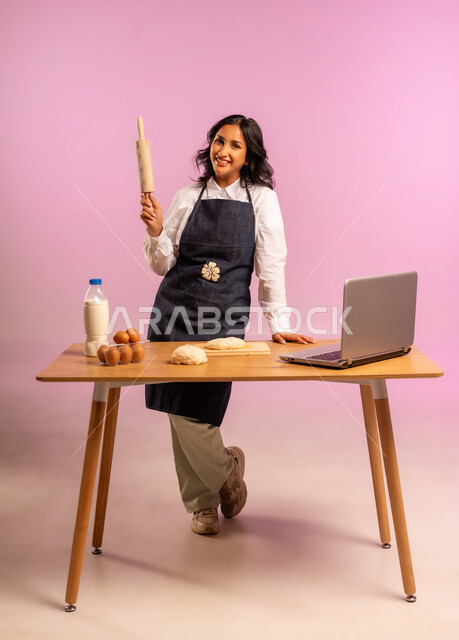 مشاركة تحضير الوصفات على مواقع التواصل الإجتماعي بإستخدام جهاز الحاسب المحمول، إستخدام أداة فرد العجين اليد (الشوبك)، بورتريه لمرأة عربية خليجية سعودية ترتدي اليونفور و تقوم بإعداد المخبوزات العربية، الجسم بالطول الكامل، خلفية ملونة