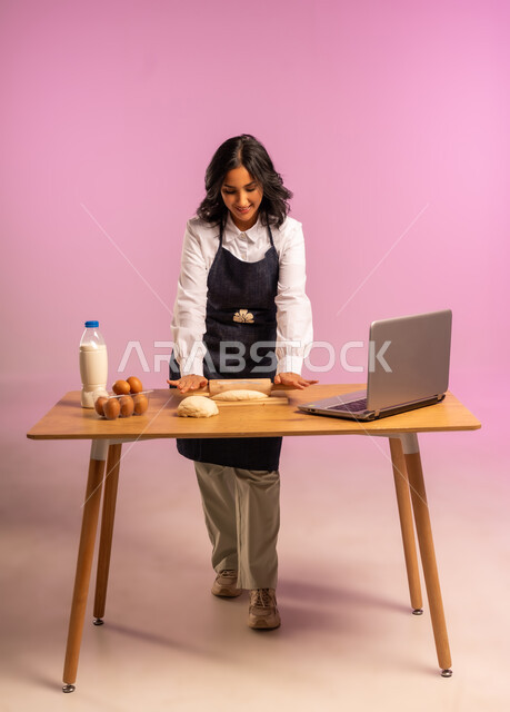 إستخدام أداة فرد العجين اليد (الشوبك)، مشاركة تحضير الوصفات على مواقع التواصل الإجتماعي بإستخدام جهاز الحاسب المحمول، بورتريه لمرأة عربية خليجية سعودية ترتدي اليونفور و تقوم بإعداد المخبوزات العربية، الجسم بالطول الكامل، خلفية ملونة