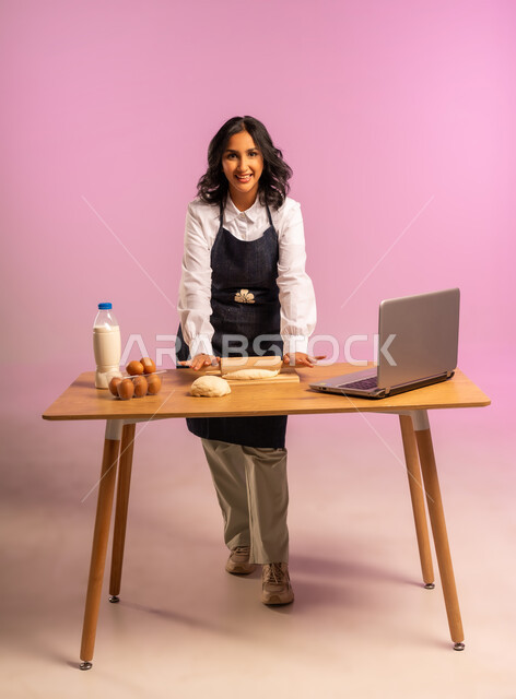 مشاركة تحضير الوصفات على مواقع التواصل الإجتماعي بإستخدام جهاز الحاسب المحمول، إستخدام أداة فرد العجين اليد (الشوبك)، بورتريه لمرأة عربية خليجية سعودية ترتدي اليونفور و تقوم بإعداد المخبوزات العربية، الجسم بالطول الكامل، خلفية ملونة
