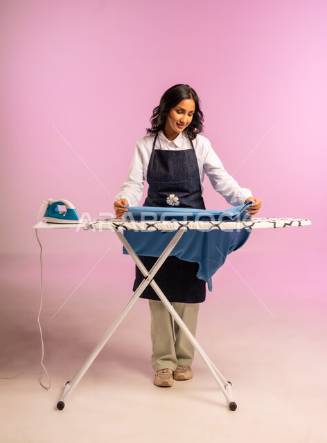 الإستعداد لكي الملابس على اللوح، تعابير تدل على الإعجاب والسعادة، بورتريه لمرأة عربية خليجية سعودية ترتدي اليونفور وتحمل بيدها المكواة الكهربائية، المهام والأعمال المنزلية الروتينية،  ترتيب وتجفيف الالبسة، الجسم بالطول الكامل، خلفية ملونة