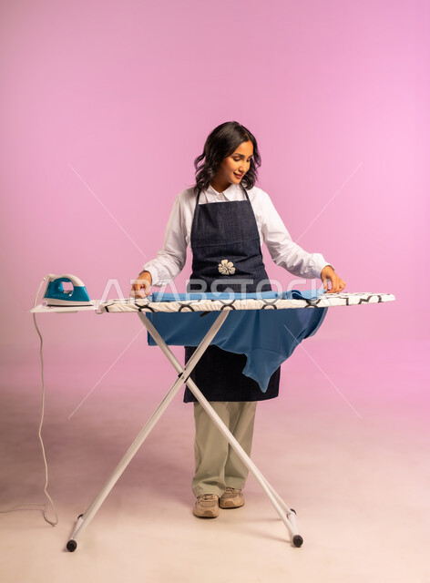 انجاز المهام والأعمال الروتينية، الاستعداد لكي الملابس على اللوح، ايماءات السعادة والسرور، بورتريه لامرأة عربية خليجية سعودية ترتدي المريول وتحمل بيدها الملابس وبجانبها المكواة الكهربائية، ترتيب وتجفيف الالبسة، الجسم بالطول الكامل، خلفية ملونة