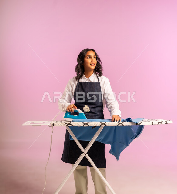 الإستعداد لكي الملابس على اللوح، تعابير تدل على الإعجاب والسعادة، بورتريه لمرأة عربية خليجية سعودية ترتدي اليونفور وتحمل بيدها المكواة الكهربائية، المهام والأعمال المنزلية الروتينية،  ترتيب وتجفيف الالبسة، الجسم بالطول الكامل، خلفية ملونة