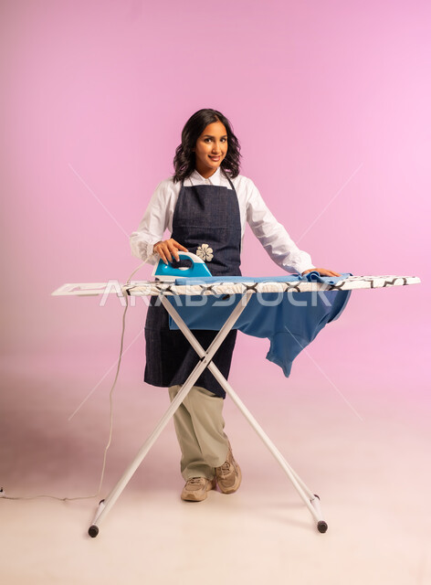 انجاز المهام والأعمال الروتينية، الاستعداد لكي الملابس على اللوح، ايماءات السعادة والسرور، بورتريه لامرأة عربية خليجية سعودية ترتدي المريول وتحمل بيدها الملابس وبجانبها المكواة الكهربائية، ترتيب وتجفيف الالبسة، الجسم بالطول الكامل، خلفية ملونة