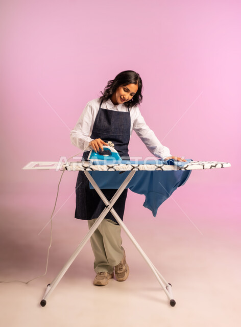 الإستعداد لكي الملابس على اللوح، تعابير تدل على الإعجاب والسعادة، بورتريه لمرأة عربية خليجية سعودية ترتدي اليونفور وتحمل بيدها المكواة الكهربائية، المهام والأعمال المنزلية الروتينية،  ترتيب وتجفيف الالبسة، الجسم بالطول الكامل، خلفية ملونة