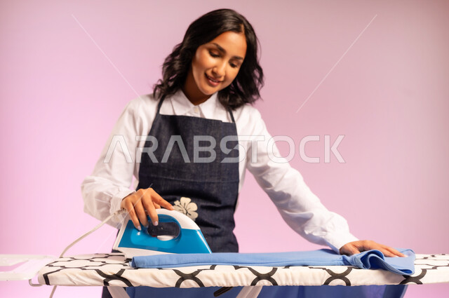Arranging and drying clothes, preparing to iron clothes on the board, expressions of admiration and happiness, close-up portrait of a Saudi Arabian Gulf woman wearing a uniform and holding an electric iron in her hand, routine household tasks and chores, colorful background