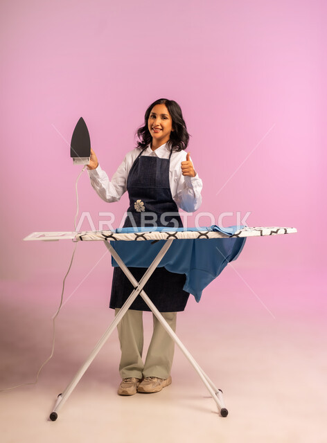 رفع اصبع الابهام بايماءات القبول والرضا، انجاز المهام والأعمال الروتينية، بورتريه لامرأة عربية خليجية سعودية ترتدي المريول تقوم بكي الملابس باستخدام المكواة، استخدام الجهاز الكهربائي، مفهوم التدبير المنزلي، تجفيف وتنظيم الملابس، الجسم بالطول الكامل، خلفية ملونة