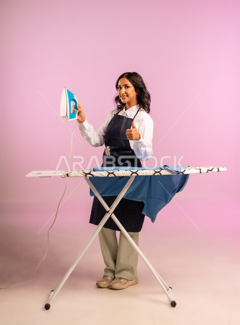 تجفيف وتنظيم الملابس، رفع اصبع الابهام بايماءات القبول والرضا، انجاز المهام والأعمال الروتينية، بورتريه لامرأة عربية خليجية سعودية ترتدي المريول تقوم بكي الملابس باستخدام المكواة، استخدام الجهاز الكهربائي، مفهوم التدبير المنزلي، الجسم بالطول الكامل، خلفية ملونة
