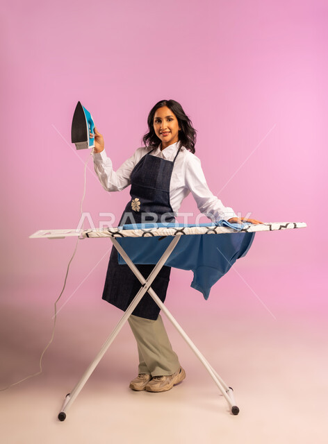الإستعداد لكي الملابس على اللوح، تعابير تدل على الإعجاب والسعادة، بورتريه لمرأة عربية خليجية سعودية ترتدي اليونفور وتحمل بيدها المكواة الكهربائية، المهام والأعمال المنزلية الروتينية،  ترتيب وتجفيف الالبسة، الجسم بالطول الكامل، خلفية ملونة