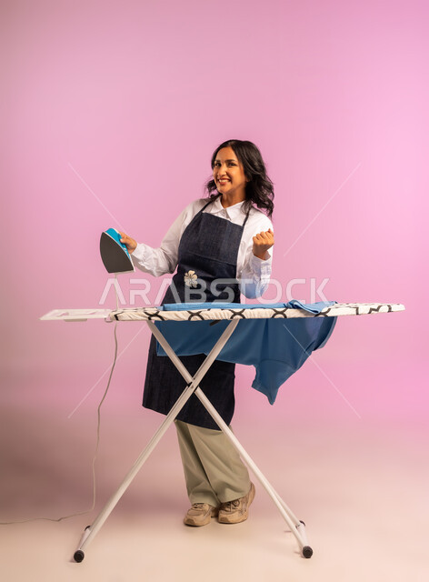 رفع قبضة اليد بايماءات الحماس والسرور، انجاز المهام والأعمال الروتينية، بورتريه لامرأة عربية خليجية سعودية مبتسمة ترتدي المريول تقوم بكي الملابس باستخدام المكواة، استخدام الجهاز الكهربائي، مفهوم التدبير المنزلي، تجفيف وتنظيم الملابس، الجسم بالطول الكامل، خلفية ملونة