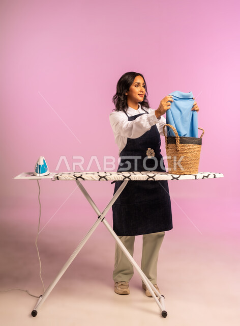 الإستعداد لكي الملابس على اللوح، تعابير تدل على الإعجاب والسعادة، بورتريه لمرأة عربية خليجية سعودية ترتدي اليونفور وتحمل بيدها المكواة الكهربائية، المهام والأعمال المنزلية الروتينية،  ترتيب وتجفيف الالبسة، الجسم بالطول الكامل، خلفية ملونة