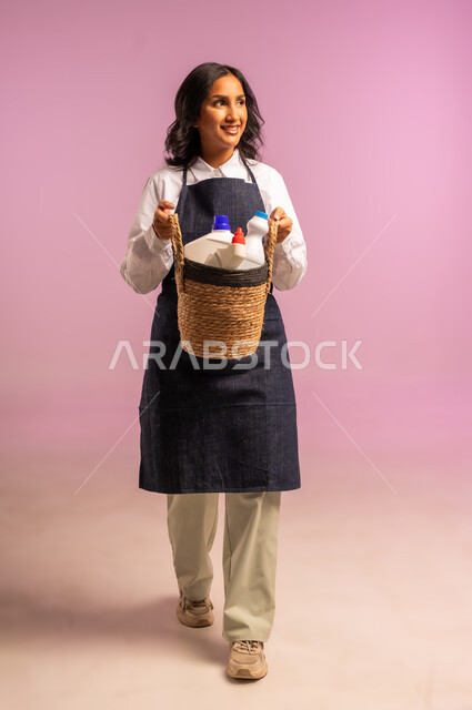Looking at a place with gestures of happiness and pleasure, tasks and routine chores, portrait of a Saudi Arabian Gulf woman wearing an apron and gloves holding a straw basket full of cleaning tools, taking care of household matters, full body, colorful background