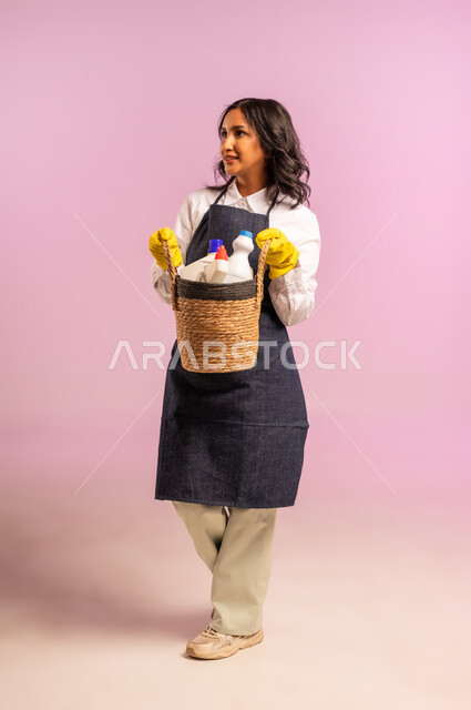 النظر الى مكان ما بايماءات السعادة والسرور، المهام والأعمال الروتينية، بورتريه لامراة عربية خليجية سعودية ترتدي المريول والقفازات تحمل بيدها سلة قش مليئة بأدوات التنظيف، الاهتمام بالأمور المنزلية، الجسم بالطول الكامل، خلفية ملونة