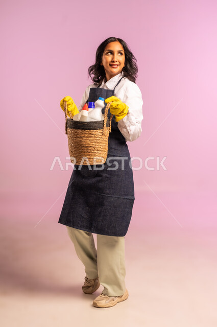 النظر الى مكان ما بايماءات السعادة والسرور، المهام والأعمال الروتينية، بورتريه لامراة عربية خليجية سعودية ترتدي المريول والقفازات تحمل بيدها سلة قش مليئة بأدوات التنظيف، الاهتمام بالأمور المنزلية، الجسم بالطول الكامل، خلفية ملونة