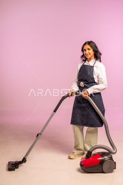أداة التنظيف الكهربائية، المهام والأعمال الروتينية، استخدام المكنسة الكهربائية، بورتريه لامرأة عربية خليجية سعودية مبتسمة ترتدي المريول تنظر إلى الكاميرا بايماءات الحماس والسرور، الاهتمام بالعمل المنزلي، الجسم بالطول الكامل، خلفية ملونة