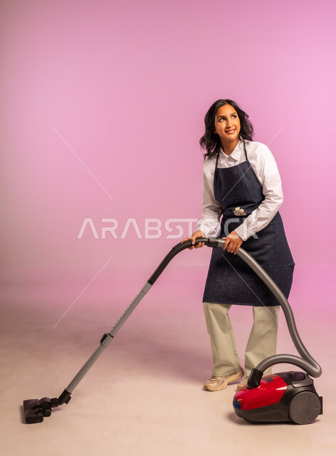 أداة التنظيف الكهربائية، المهام والأعمال الروتينية، استخدام المكنسة الكهربائية، بورتريه لامرأة عربية خليجية سعودية مبتسمة ترتدي المريول تنظر إلى مكان ما بايماءات الحماس والسرور، الاهتمام بالعمل المنزلي، الجسم بالطول الكامل، خلفية ملونة
