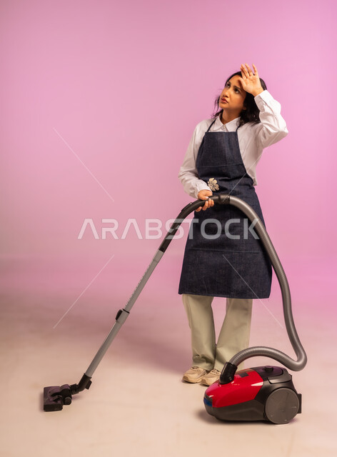 استخدام المكنسة الكهربائية، بورتريه لامرأة عربية خليجية سعودية ترتدي المريول تضع يدها على رأسها بايماءات التعب والارهاق، المهام والأعمال الروتينية، أداة التنظيف الكهربائية، الاهتمام بالعمل المنزلي، الجسم بالطول الكامل، خلفية ملونة