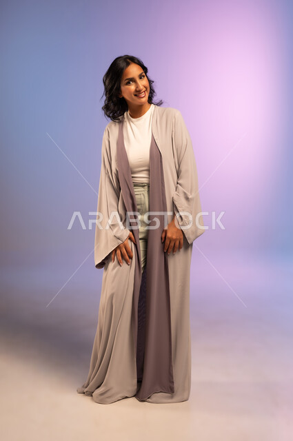 الثقة والاعتزاز بالنفس، الاهتمام بالأناقة والمظهر الخارجي، مفهوم الأنوثة والنعومة، بورتريه لامرأة عربية خليجية سعودية مبتسمة  ترتدي عباءة و تنظر الى شيء ما بإيماءات السرور، الجسم بالطول الكامل، خلفية ملونة
