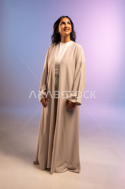 Concern for elegance and appearance, concept of femininity and softness, confidence and self-esteem, portrait of a smiling Saudi Arabian Gulf woman wearing an abaya and looking at something with gestures of pleasure, full-length body, colored background
