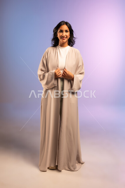 The concept of femininity and softness, confidence and self-esteem, interest in elegance and appearance, portrait of a smiling Saudi Arabian Gulf woman wearing an abaya and looking at the camera with gestures of pleasure, full-length body, colored background