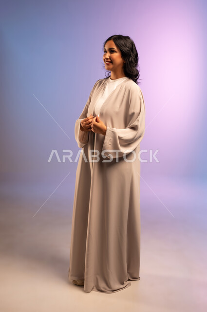Concern for elegance and appearance, concept of femininity and softness, confidence and self-esteem, portrait of a smiling Saudi Arabian Gulf woman wearing an abaya and looking at something with gestures of pleasure, full-length body, colored background