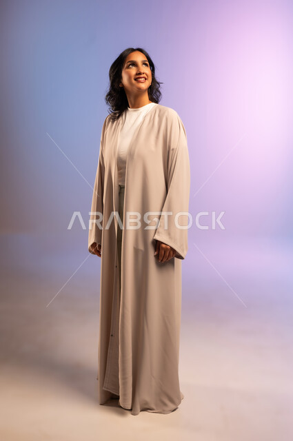 Concern for elegance and appearance, concept of femininity and softness, confidence and self-esteem, portrait of a smiling Saudi Arabian Gulf woman wearing an abaya and looking at something with gestures of pleasure, full-length body, colored background