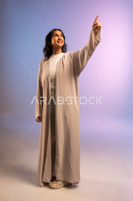 الثقة والاعتزاز بالنفس، الاهتمام بالأناقة والمظهر الخارجي، مفهوم الأنوثة والنعومة، بورتريه لامرأة عربية خليجية سعودية مبتسمة ترتدي العباءة تشير باصبع السبابة الى شيء ما بإيماءات السعادة والسرور، الجسم بالطول الكامل، خلفية ملونة
