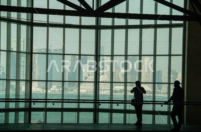 Modern architectural art, decoration and interior design of the Museum of Islamic Art in Qatar, silhouette from behind of two men standing in front of the huge glass facade overlooking the Arabian Sea, a famous tourist place and landmark in Doha